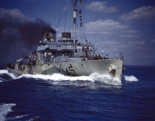 Colour photograph of a Royal Canadian Navy corvette under way at top speed. Thick black smoke pours from a funnel. Number K145 is written in black on the grey vessel. 